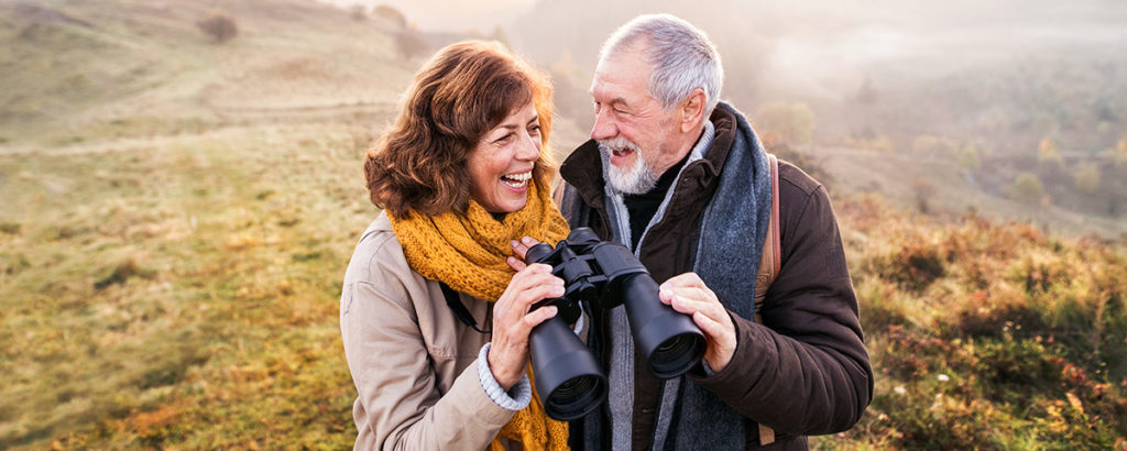 Smiling couple outdoors holding binoculars surrounded by hills.