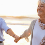 Happy, smiling 50+ couple holding hands and walking along the beach together with the sunset in the background.